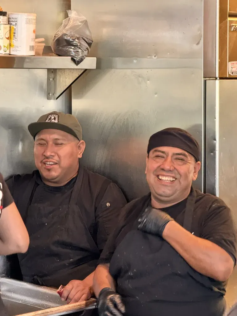 Two cooks laughing at the dish sink, in black aprons and gloves.